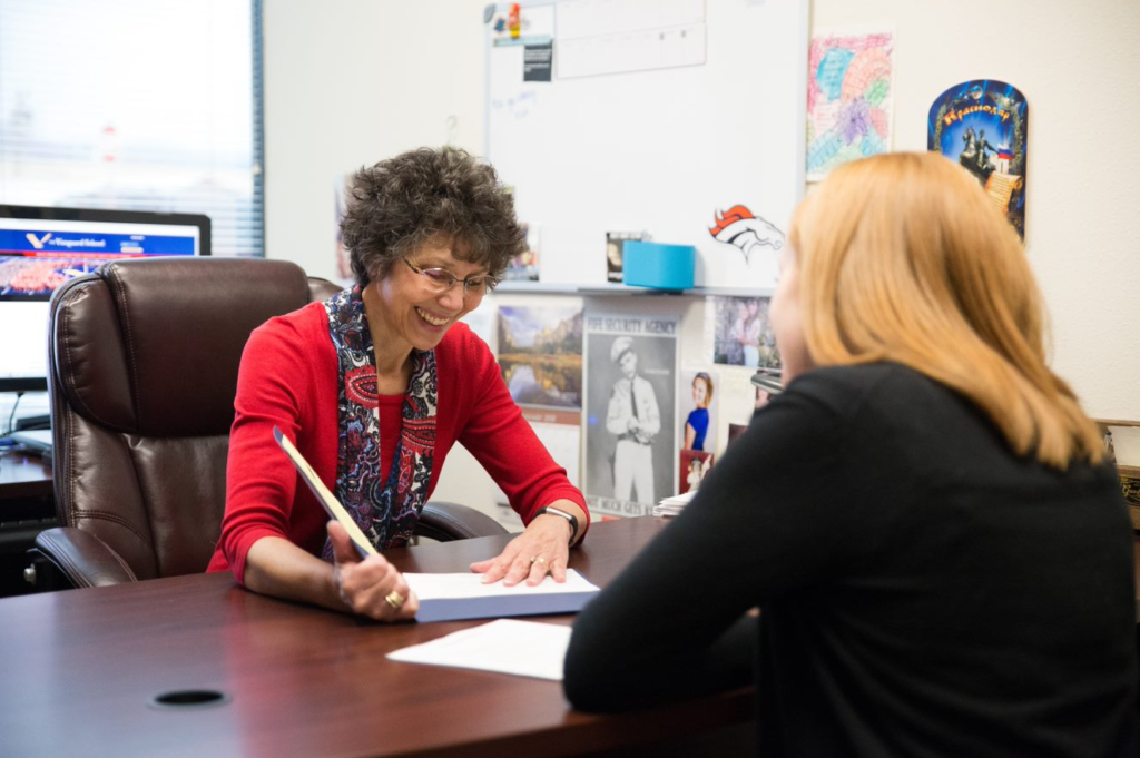 A school counselor shows some papers inside a blue folder to the student with their back to the camera across a desk